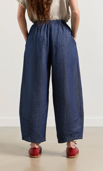 Plümo- Person stands with hands in pockets, wearing Mara Barrel Pants in indigo and red shoes, seen from the back.