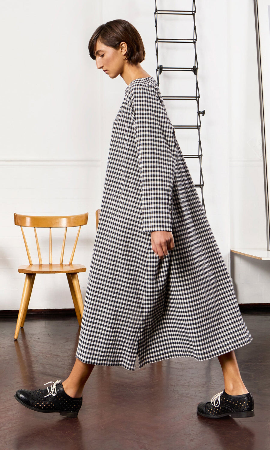 Plümo- Person in a Marsell Dress - Gingham walks indoors past a wooden chair on a dark floor.