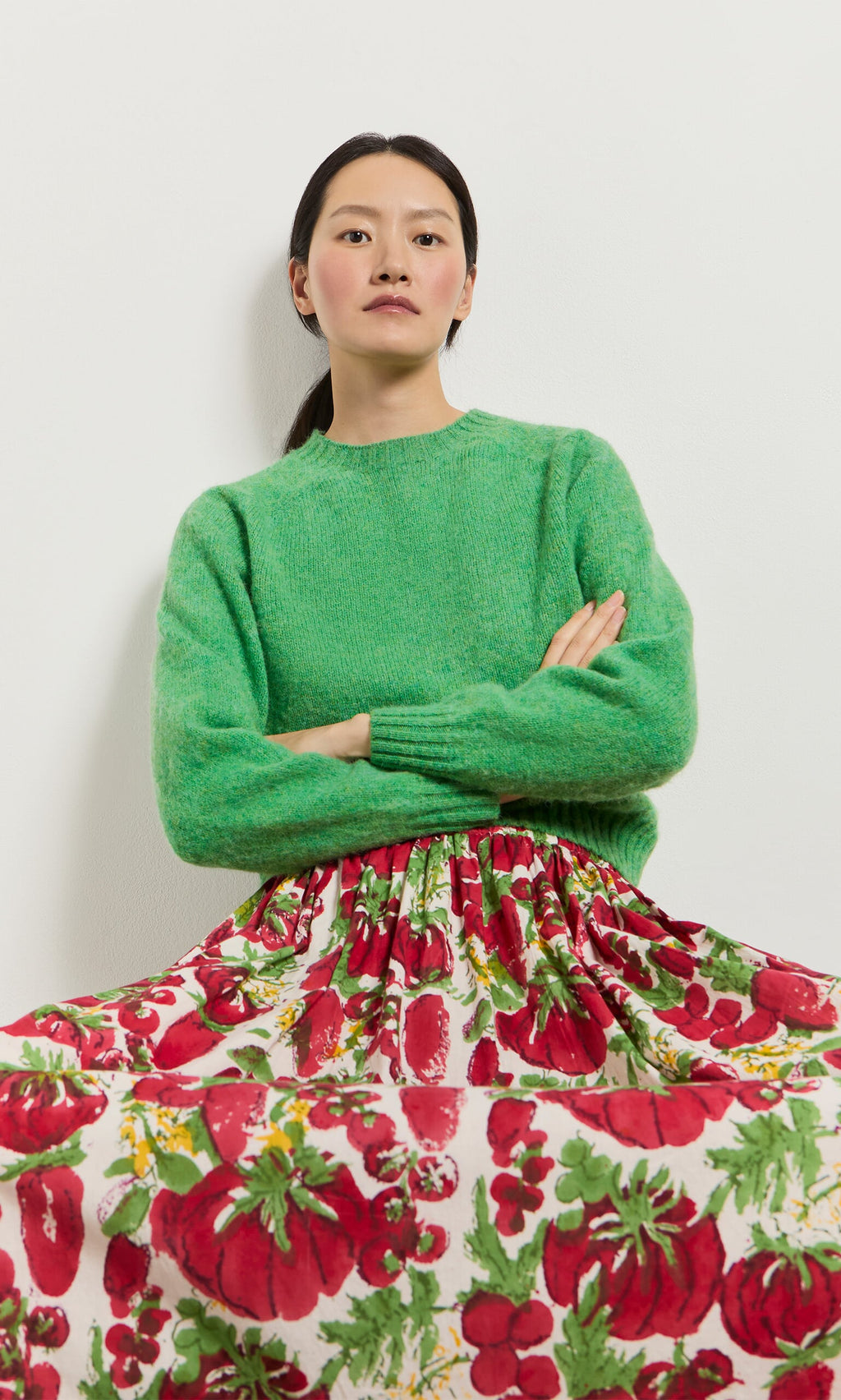 Plümo- Woman with straight dark hair in a green sweater and Marzano Skirt, sitting against a white wall.