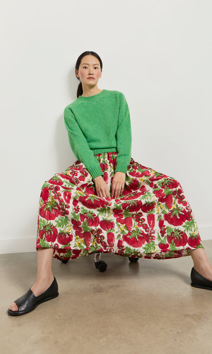 Plümo- Woman in a green sweater and Marzano Skirt with elastic waist sits on a chair, wearing black open-toe shoes.