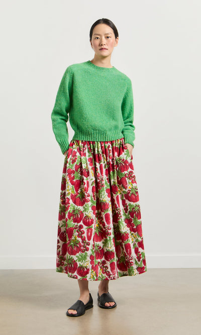 Plümo- Woman in a green sweater and Marzano Skirt with black sandals, standing against a plain background.