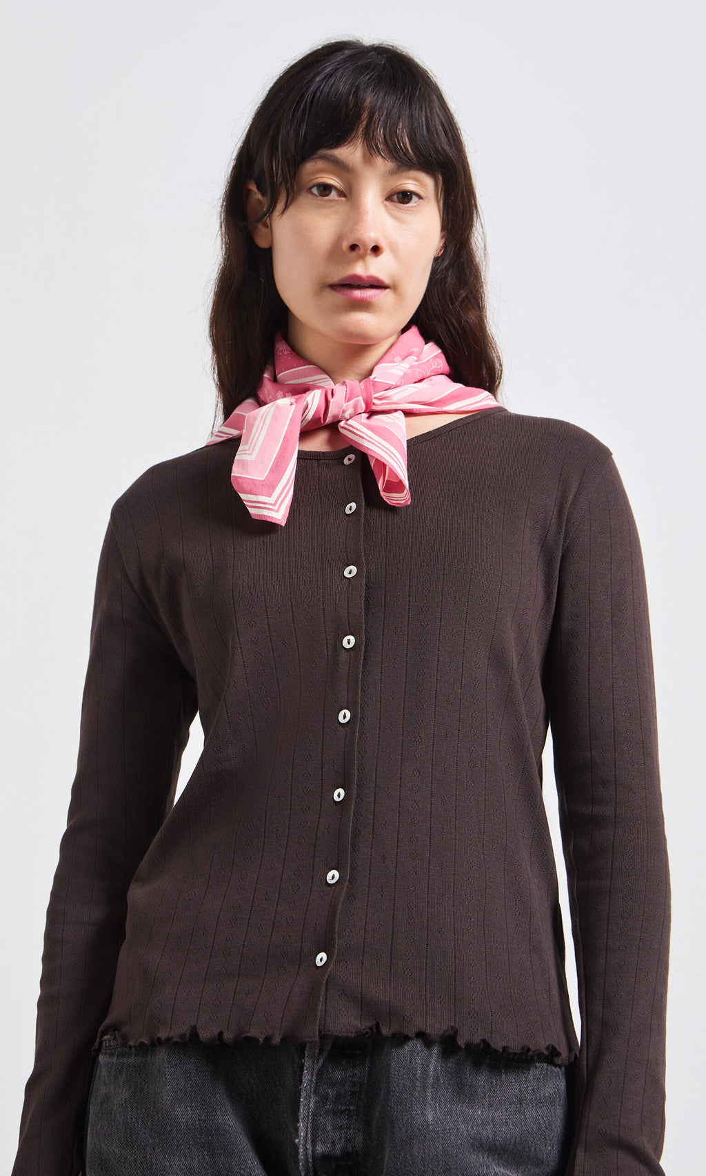 Plümo- Woman with long dark hair wears the Eddie Cardigan, black jeans, and a pink striped scarf.