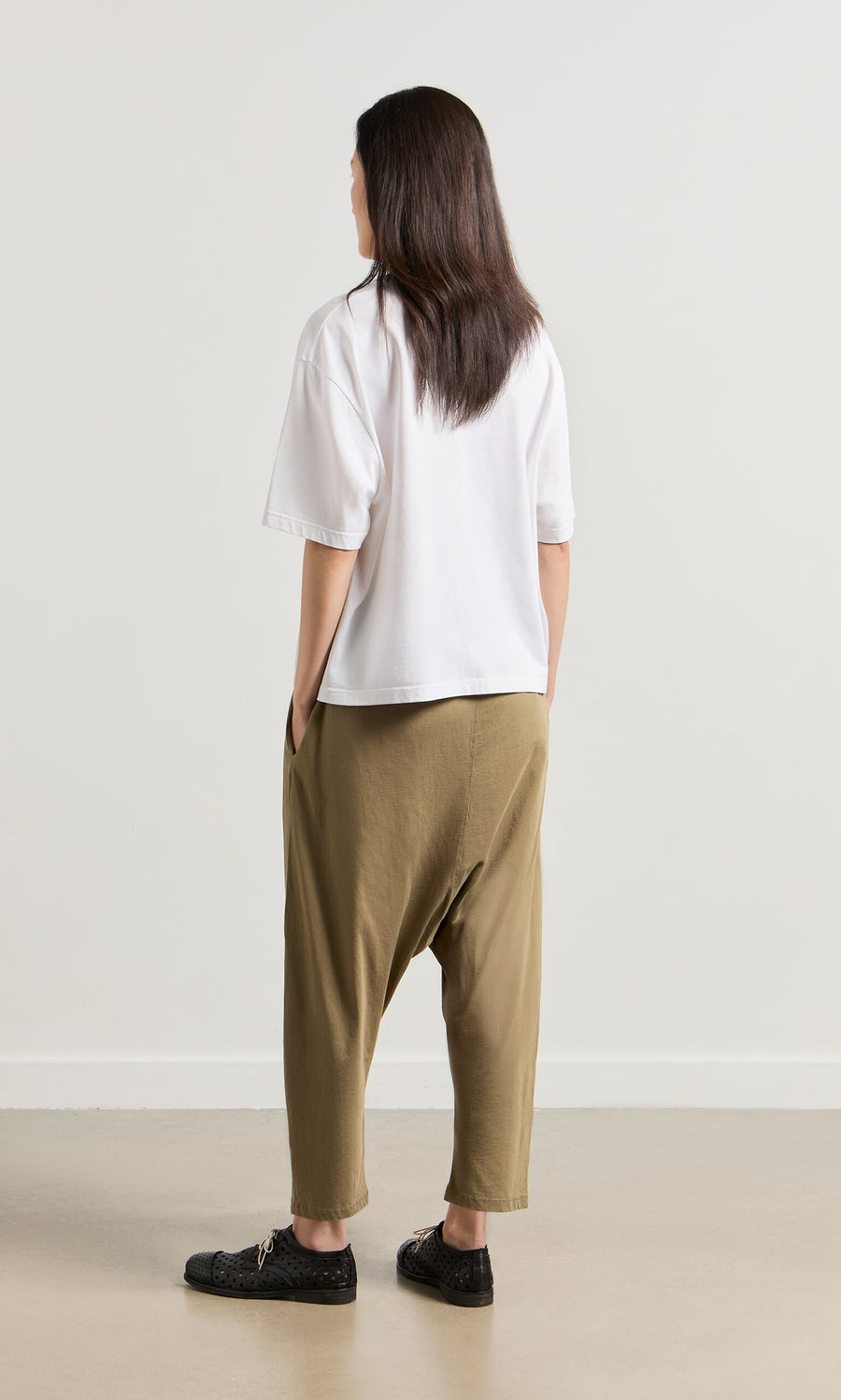 Plümo- Person stands indoors facing away, in a white shirt, black shoes, and Mirana Pants on a light floor.