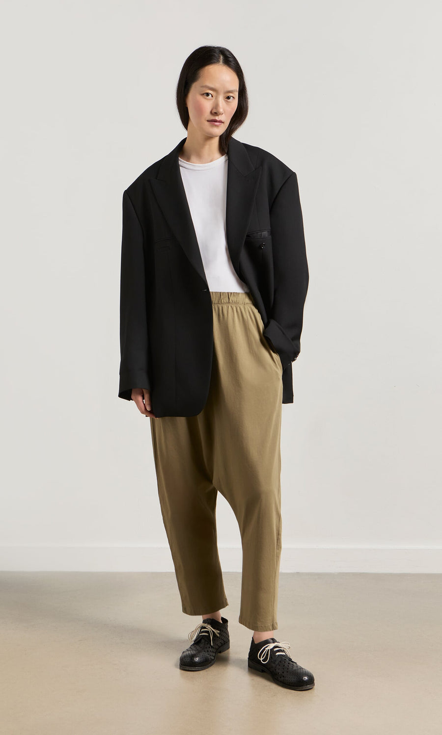 Plümo- Someone in a black blazer, white tee, and Mirana Pants stands against a plain light background.
