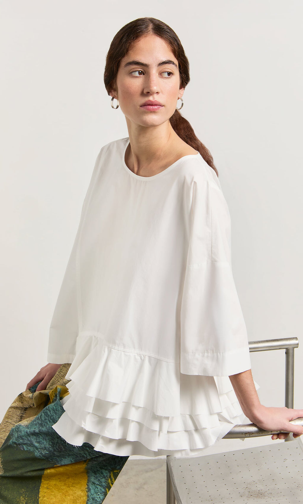 Plümo- Woman in the Mirelle Blouse, a white ruffle-hem top, sits on a metal bench gazing to the side with a neutral look.