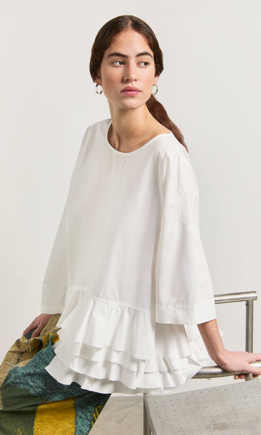 Plümo- Woman in the Mirelle Blouse, a white ruffle-hem top, sits on a metal bench gazing to the side with a neutral look.