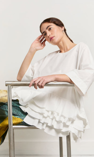 Plümo- Woman in the Mirelle Blouse with a ruffle hem sits thoughtfully on a metal chair, head resting on her hand.