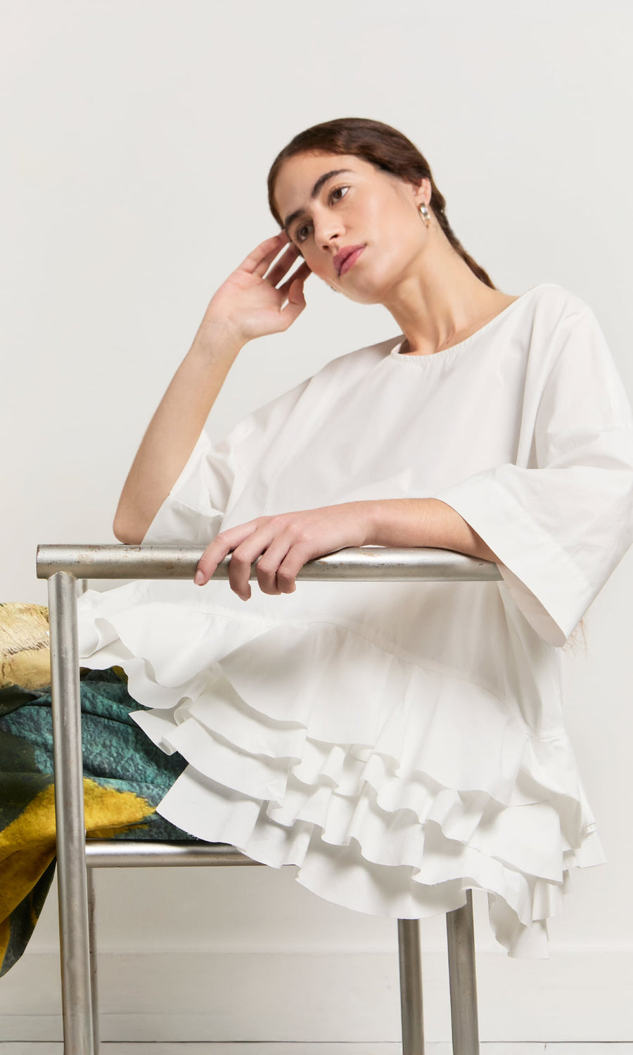 Plümo- Woman in the Mirelle Blouse with a ruffle hem sits thoughtfully on a metal chair, head resting on her hand.