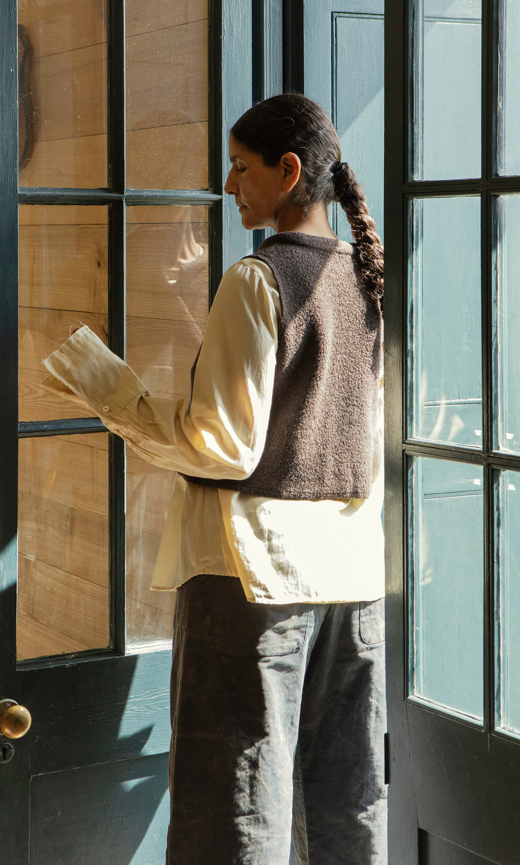 Plümo  Person with long braid in a Mons Felted Waistcoat stands by glass doors as sunlight streams through window panes.