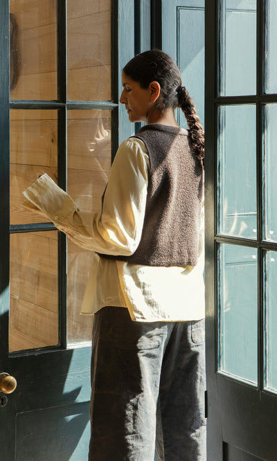 Plümo  Person with long braid in a Mons Felted Waistcoat stands by glass doors as sunlight streams through window panes.