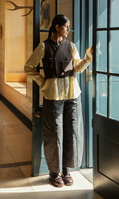 Plümo  Someone in loose pants and layered top, wearing the Mons Felted Waistcoat, stands by a glass door, looking out.