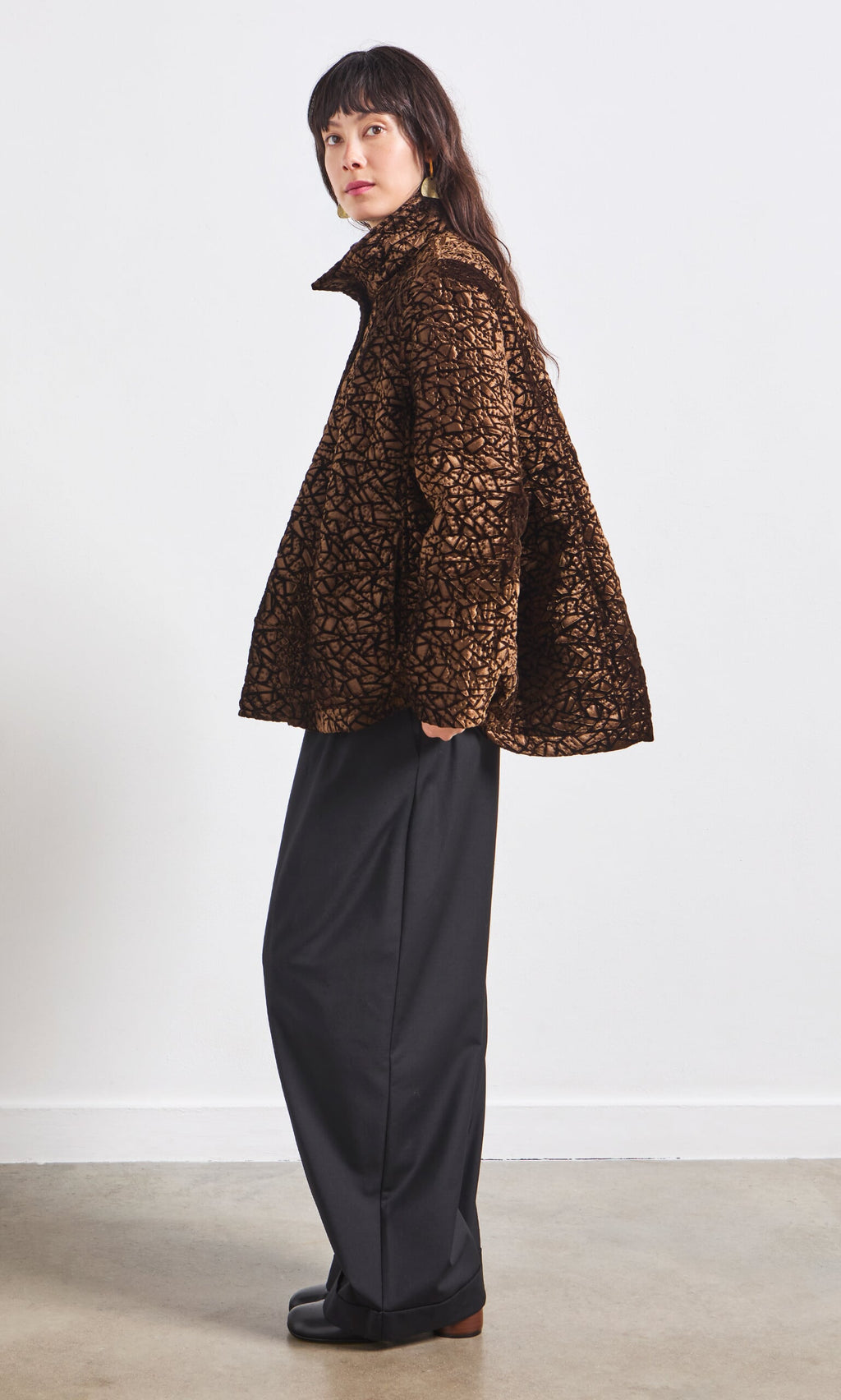 Plümo- Woman in a Murano Jacket with a geometric pattern and wide-leg black pants stands sideways against a white wall.
