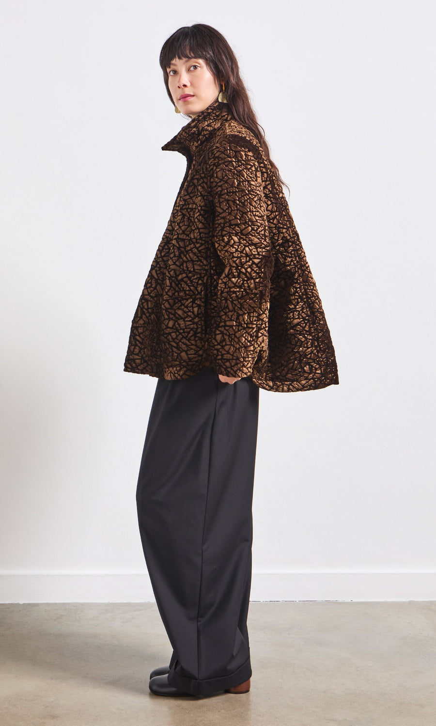 Plümo- Woman in a Murano Jacket with a geometric pattern and wide-leg black pants stands sideways against a white wall.