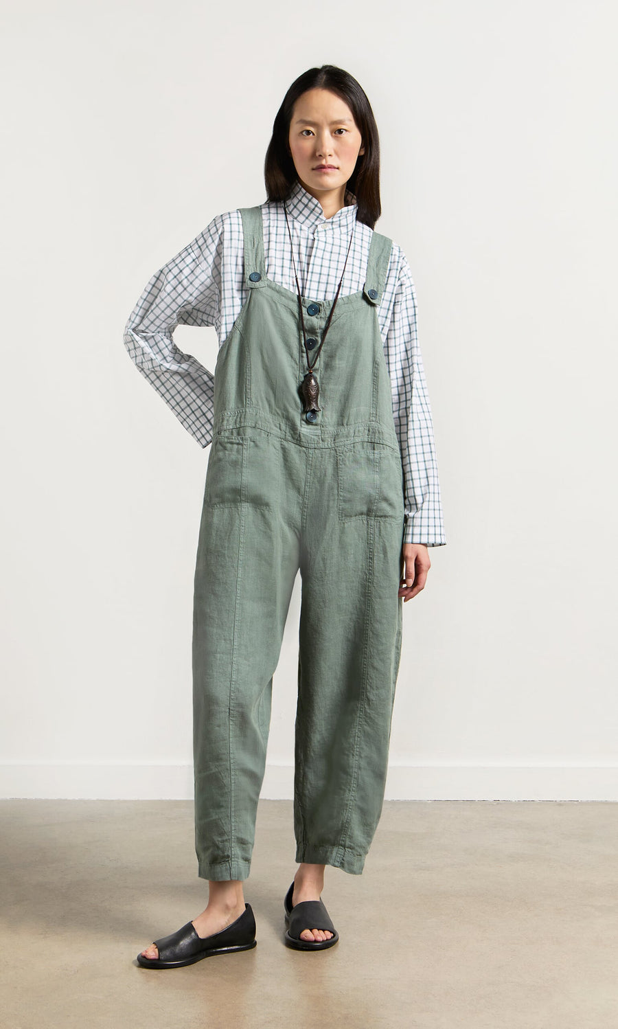 Plümo- Person standing against a white wall wearing Myrtle Linen Dungarees, a plaid shirt, black sandals, and a long necklace.