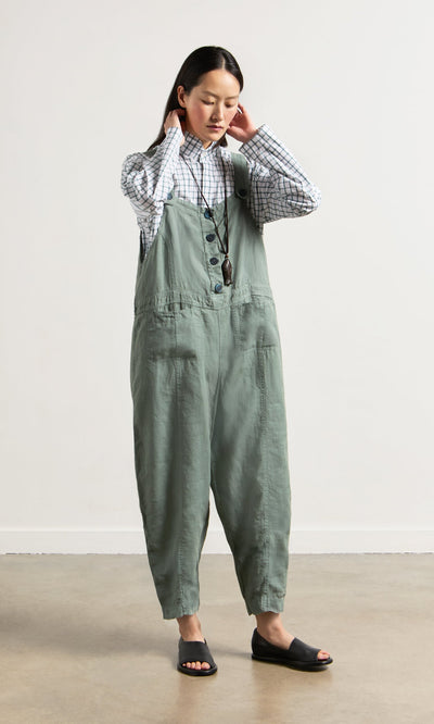 Plümo- Woman in a checked shirt and Myrtle Linen Dungarees stands indoors, touching her neck and looking down.