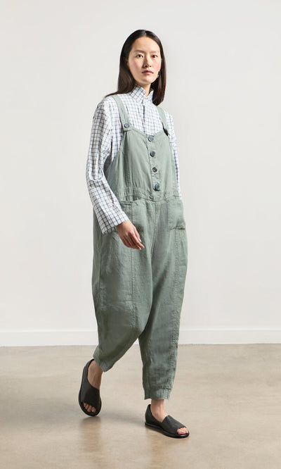 Plümo- A woman stands indoors on a light floor, wearing Myrtle Linen Dungarees, a plaid shirt, and black open-toe sandals.