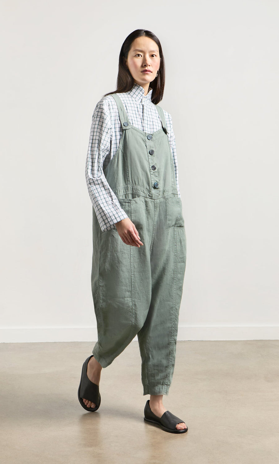 Plümo- A woman stands indoors on a light floor, wearing Myrtle Linen Dungarees, a plaid shirt, and black open-toe sandals.