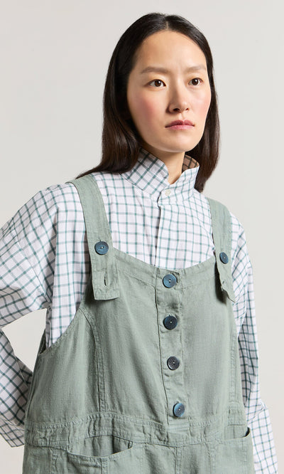 Plümo- Woman in Myrtle Linen Dungarees over a white and green checkered shirt, looking slightly to the side.