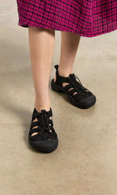 Plümo- Person wearing black Newport Sandals with arch support and a pink plaid skirt on a light concrete floor.