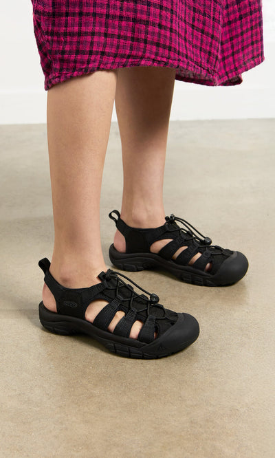 Plümo- Person in black Newport Sandals and a pink plaid skirt stands on a light-colored floor.