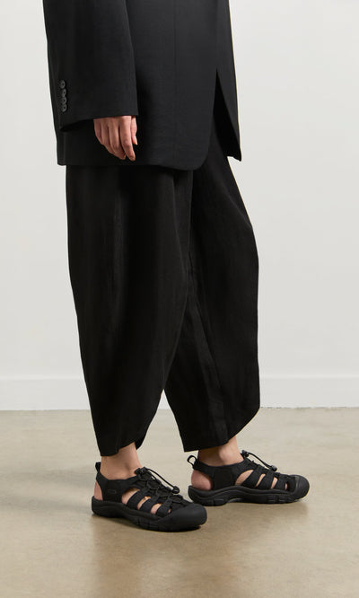 Plümo- A person in loose black pants, a black blazer, and Newport Sandals stands on a concrete floor by a white wall.