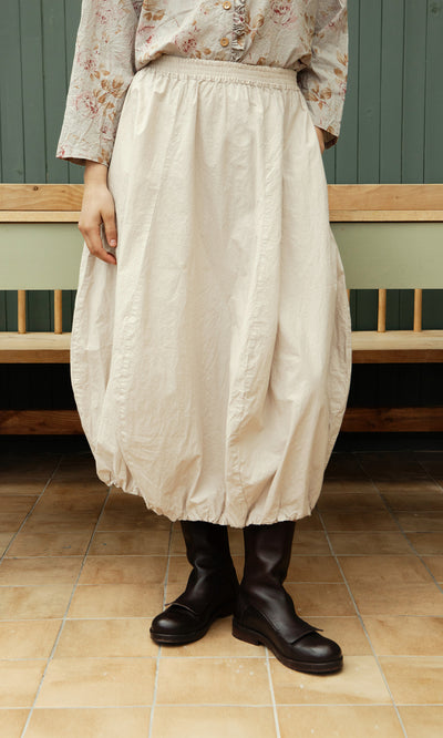Plümo  Person in the Edina Skirt - Nougat, floral blouse, and black boots stands on tiled floor by a wooden bench.
