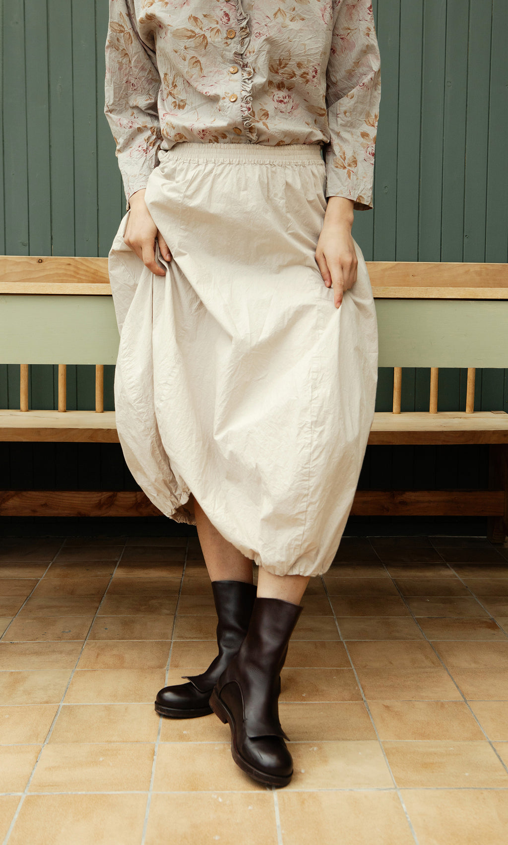 Plümo  Person in a floral blouse, Edina Skirt - Nougat, and black boots stands on tan tiles by a wooden bench.