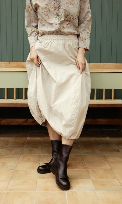 Plümo  Person in a floral blouse, Edina Skirt - Nougat, and black boots stands on tan tiles by a wooden bench.