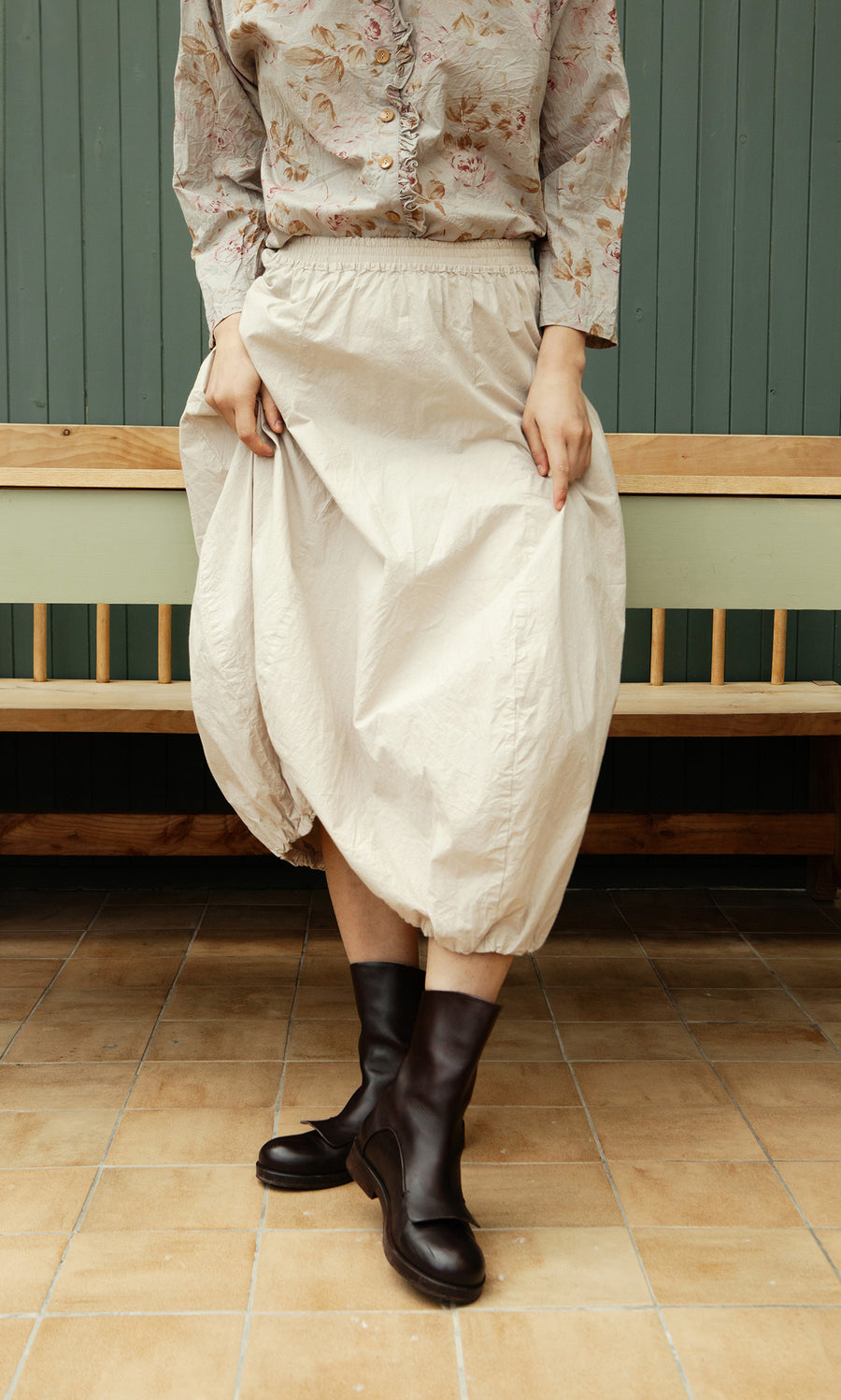 Plümo  Person in a floral blouse, Edina Skirt - Nougat, and black boots stands on tan tiles by a wooden bench.