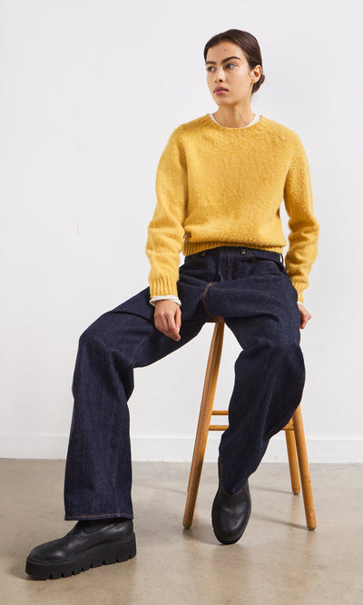 Plümo- Person in a yellow sweater and Japanese Raw Denim Jeans sits on a wooden stool against a plain white wall.