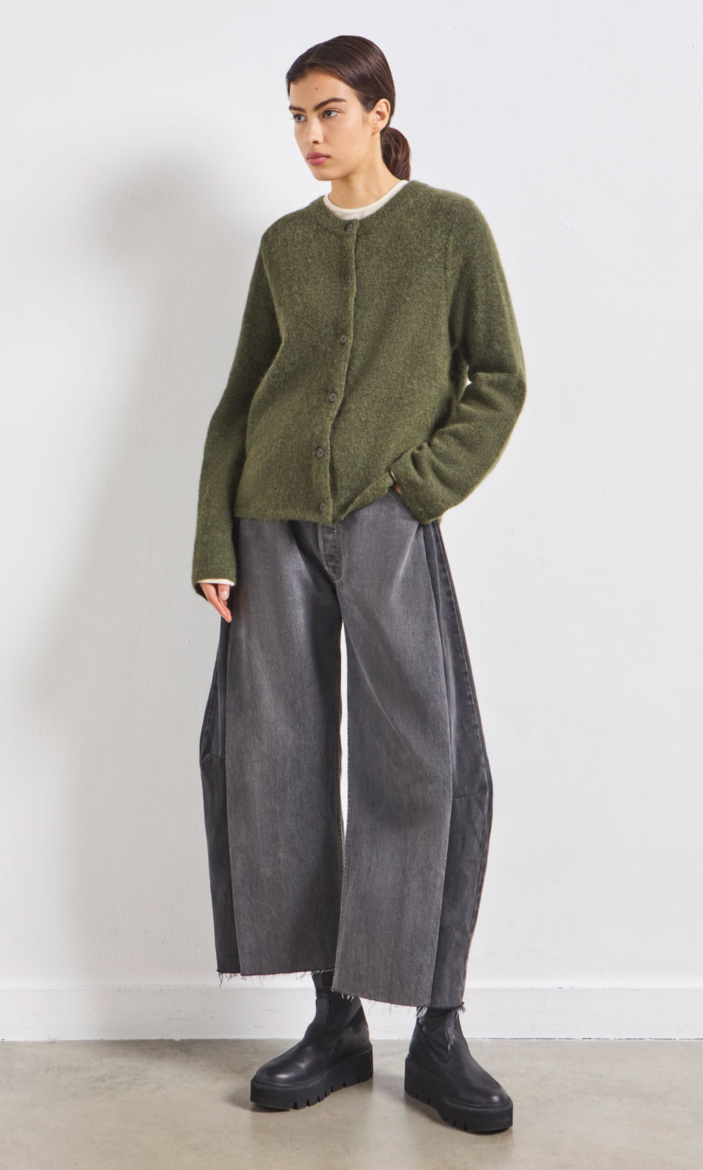 Plümo- Person wears an olive green cardigan, wide-leg gray jeans, and black boots against a white wall.