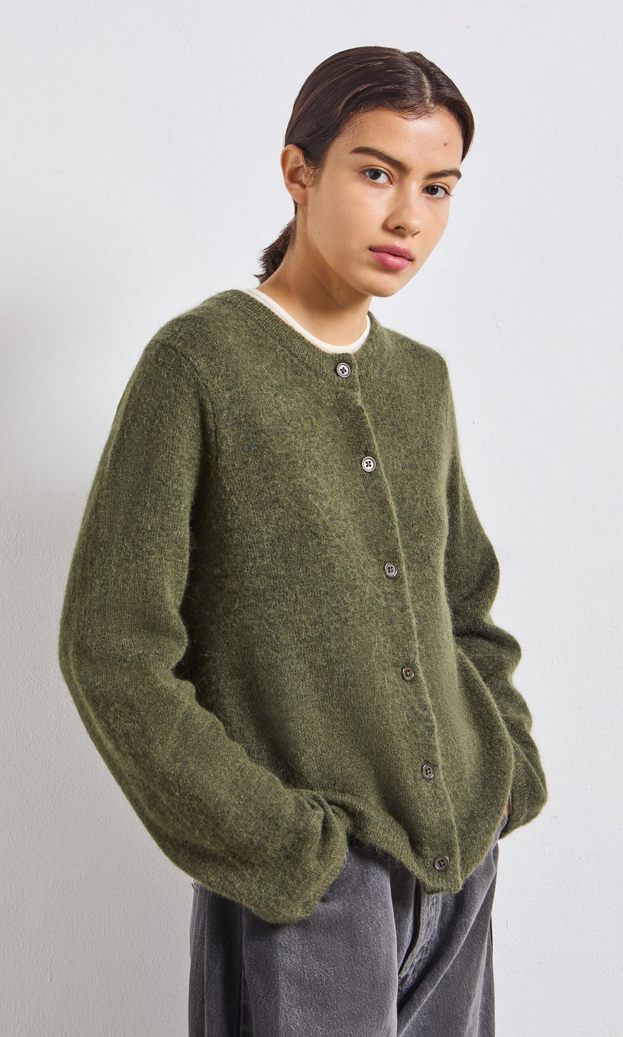 Plümo- Person wearing an olive green cardigan and grey pants, standing against a plain white background.