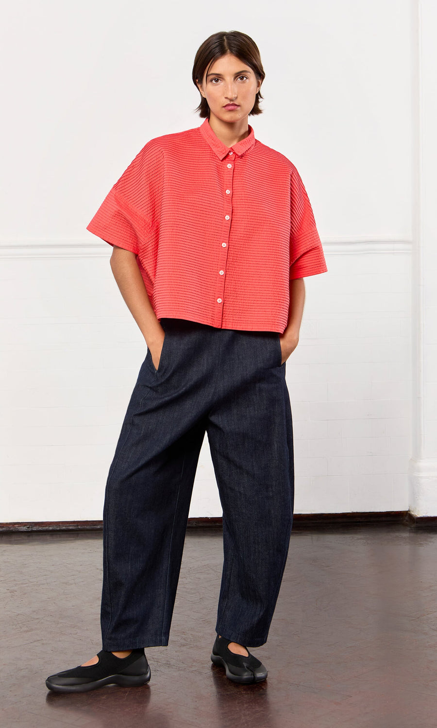 Plümo- Person in a loose red short-sleeve shirt, Lugo Barrel Jeans, and black shoes with hands in pockets indoors.