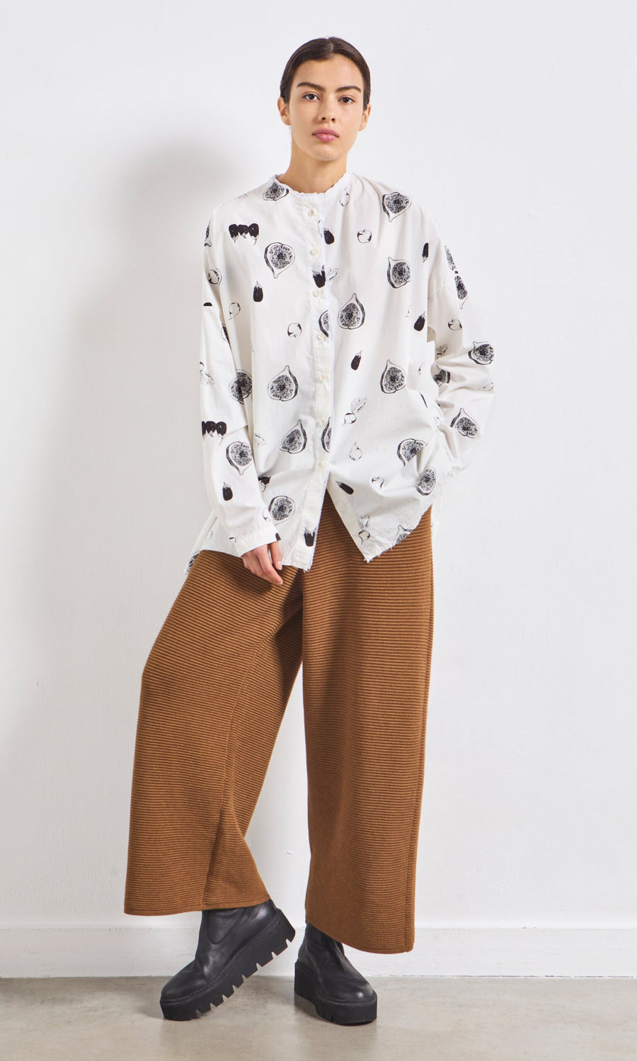 Plümo- Person wearing a white shirt, Orla Pants - Chestnut with an elastic waistband, and black platform shoes against a white wall.