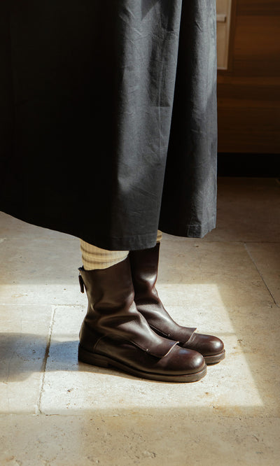 Plümo  Person in Over Boots—artisan-made Italian leather—styled with a long black skirt and cream socks on stone floor.