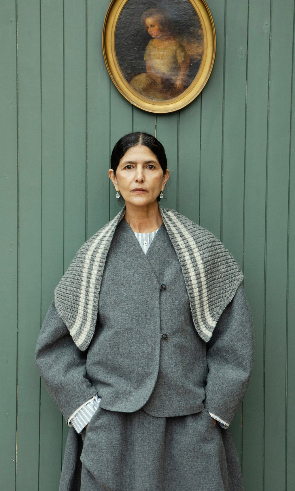 Plümo  Woman in gray coat and Oyster Triangle Scarf stands by green wall with child’s oval portrait above her head.