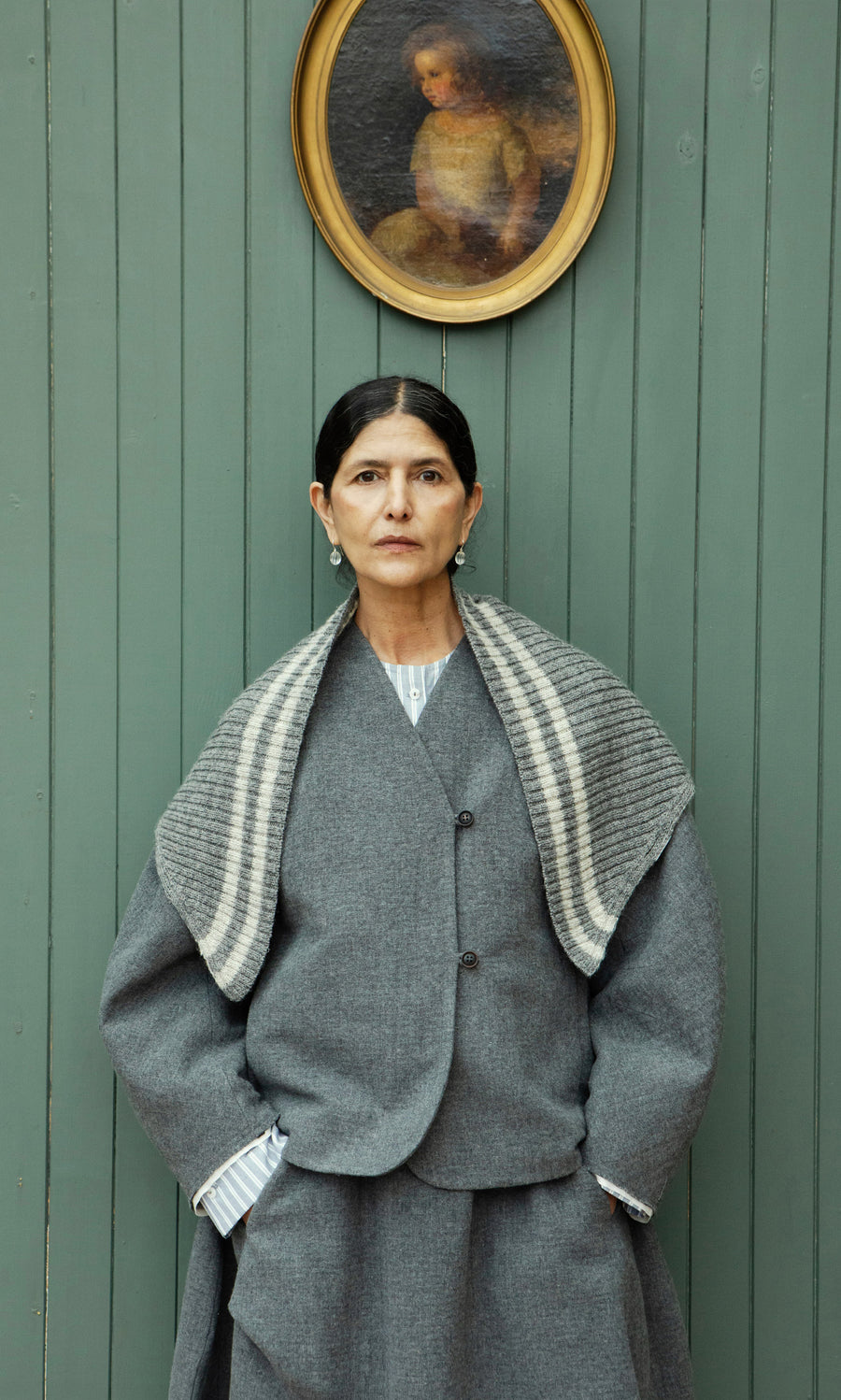 Plümo  Woman in gray coat and Oyster Triangle Scarf stands by green wall with child’s oval portrait above her head.