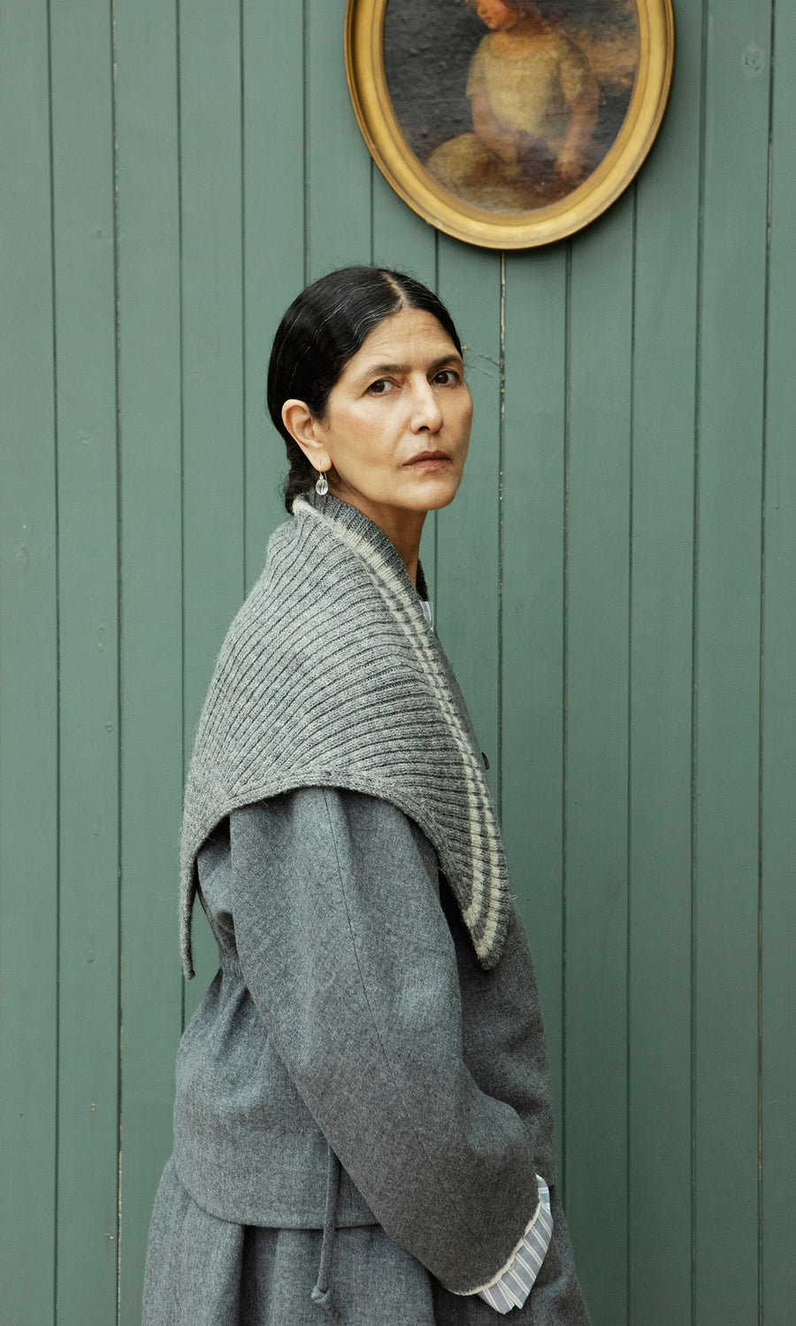 Plümo  Woman in a gray look wears the Oyster Triangle Scarf, standing sideways by a green paneled wall and framed art.