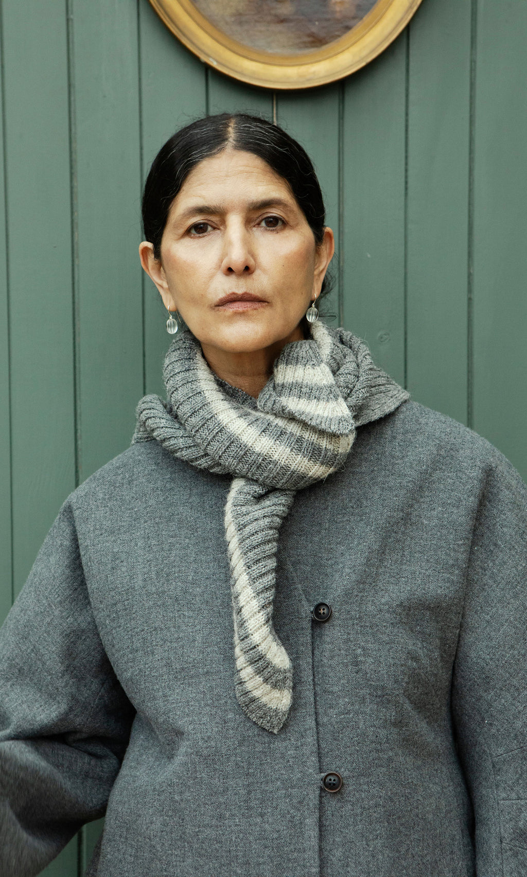 Plümo  Woman in a grey coat and Oyster Triangle Scarf stands before a green paneled wall, looking at the camera.