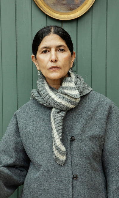 Plümo  Woman in a grey coat and Oyster Triangle Scarf stands before a green paneled wall, looking at the camera.