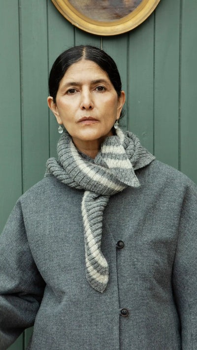 Plümo  A woman in a gray coat and Oyster Triangle Scarf stands by a green wall, looking directly at the camera.