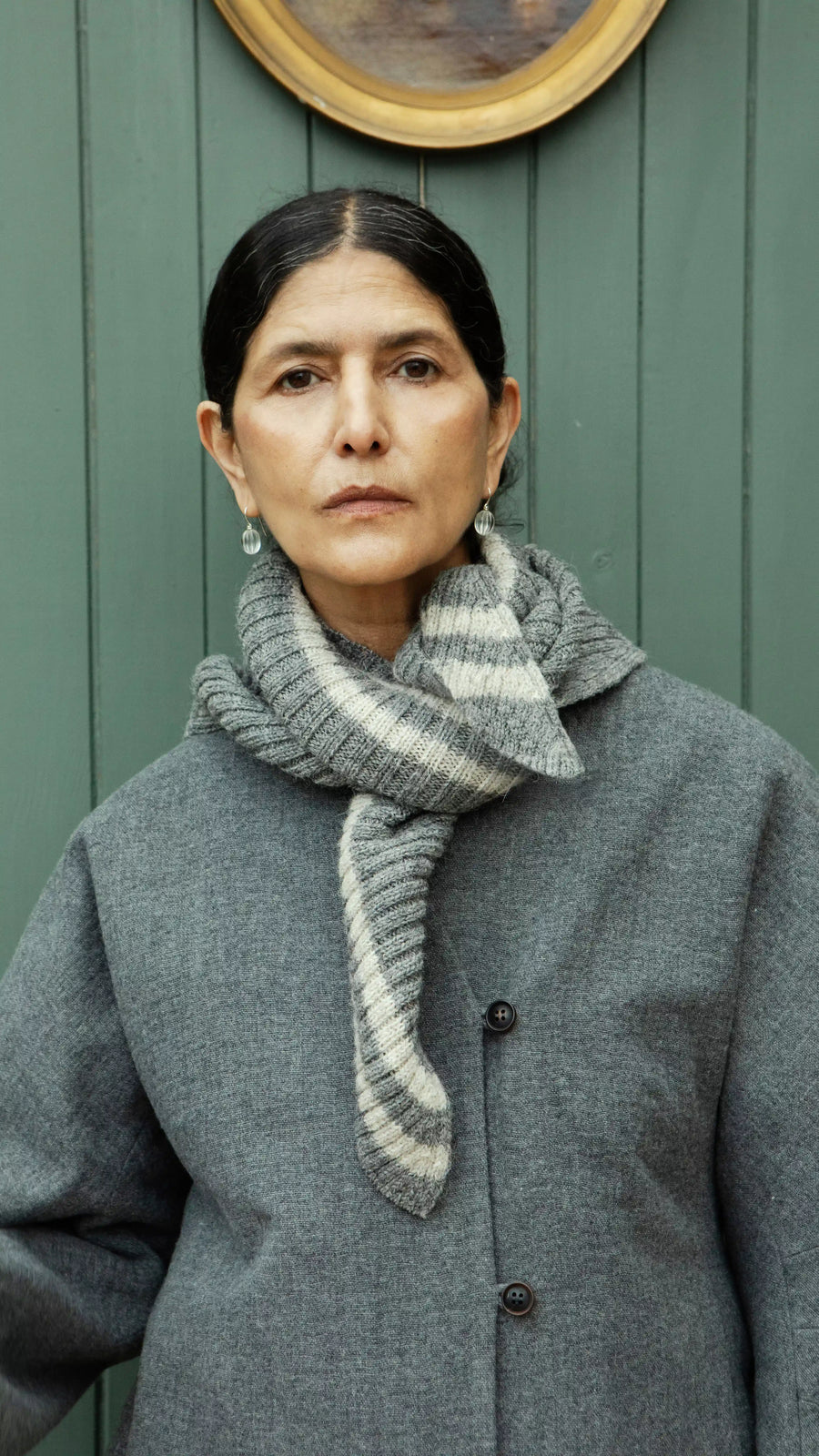 Plümo  A woman in a gray coat and Oyster Triangle Scarf stands by a green wall, looking directly at the camera.