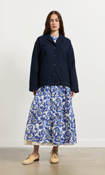 Plümo- Woman wearing the Maren Jacket over a blue and white floral dress with tan woven shoes, standing against a plain backdrop.