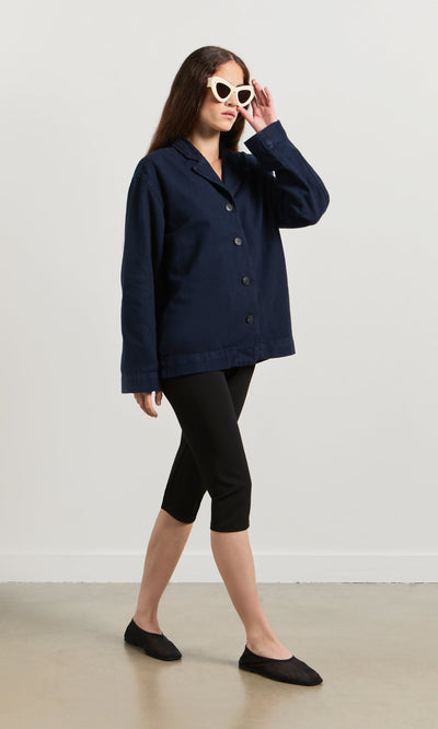 Plümo- Woman in the navy Maren Jacket, black capri pants, black flats, and white sunglasses walking indoors.