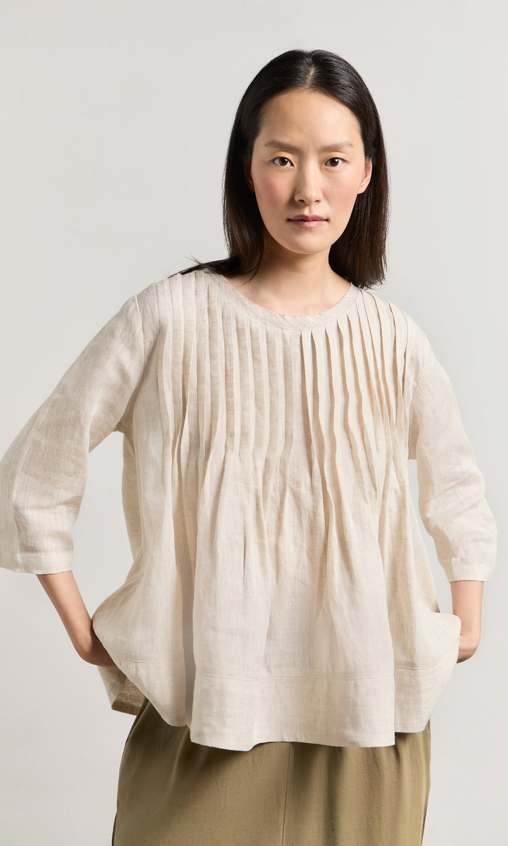 Plümo- A woman in a relaxed fit, pinch-pleated Pantry Blouse and olive pants stands hands-in-pockets, facing forward.