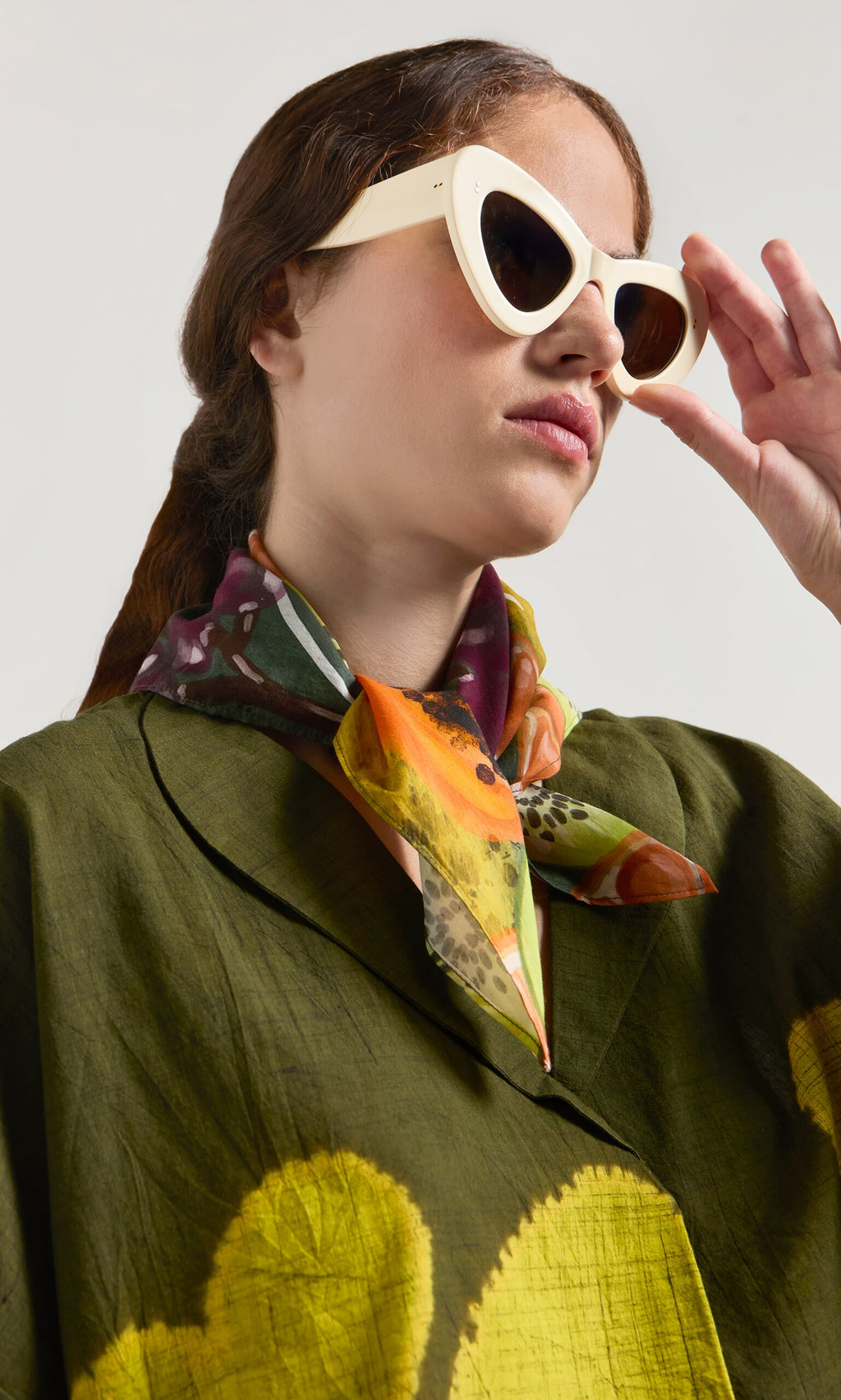 Plümo- Woman in white sunglasses, green coat, and Papaya Necktie scarf, looking upward with hand near her face.
