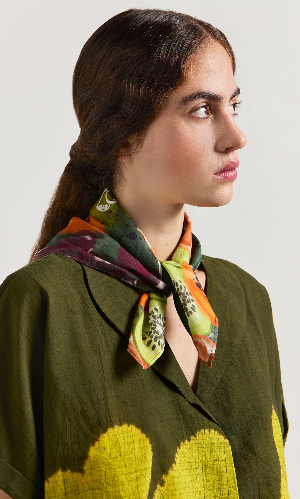 Plümo- Woman with long brown hair in a green shirt wears the ethically made Papaya Necktie scarf, looking to the side.