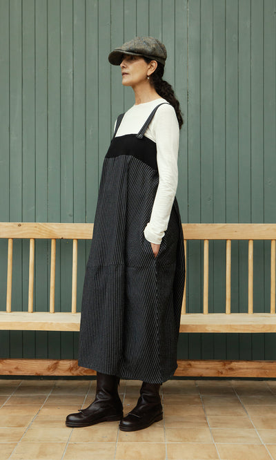 Plümo  Woman in a Pinstripe Dungaree Dress, white shirt, cap, and boots stands by a bench against a green wall.