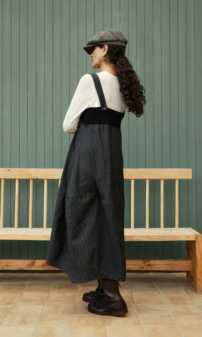 Plümo  Woman in a pinstripe dungaree dress, cream top, boots, and cap stands sideways by a bench.
