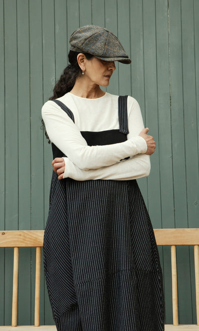 Plümo  Woman in a Pinstripe Dungaree Dress and flat cap stands with arms crossed by a green paneled wall.